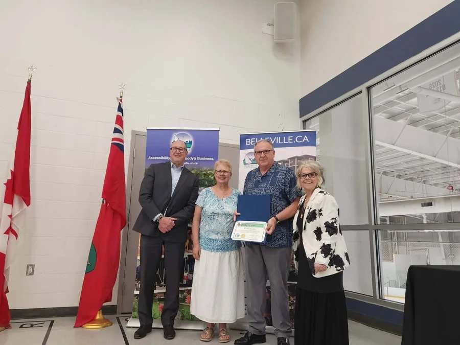 Steve Westlake accepting the City of Belleville 2026 Accessibility Excellence Award, holding the framed certificate in front of the city's accessibility banners and the Canadian and Ontario flags
